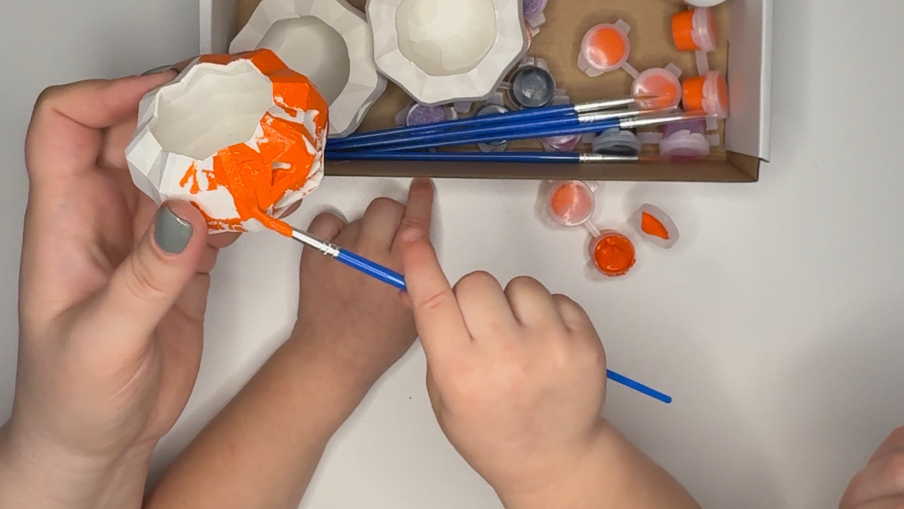 Child painting a plaster item with adult supervision, surrounded by art supplies.